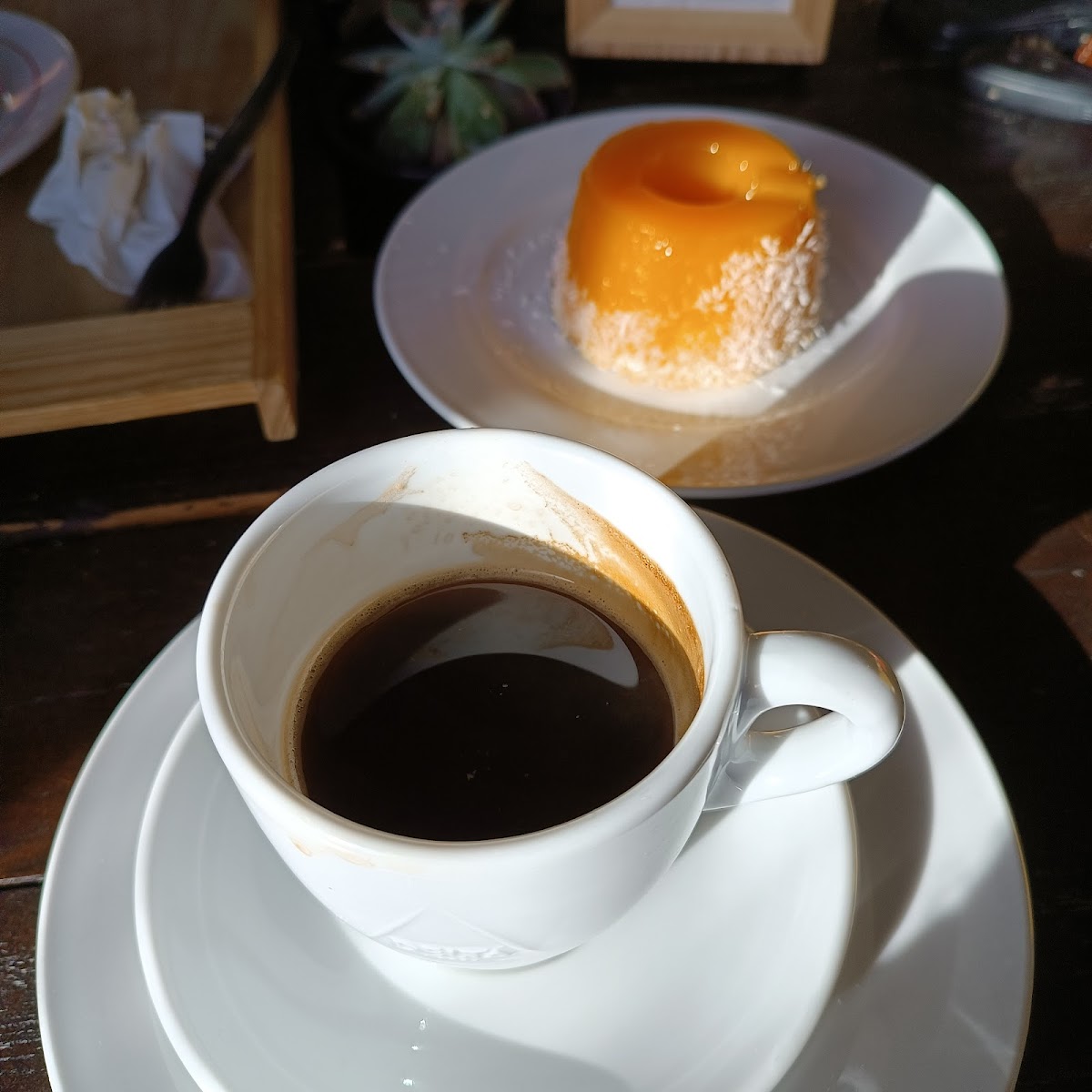 Chávena de café preto e, ao fundo, um quindim de laranja, à luz quente da manhã.