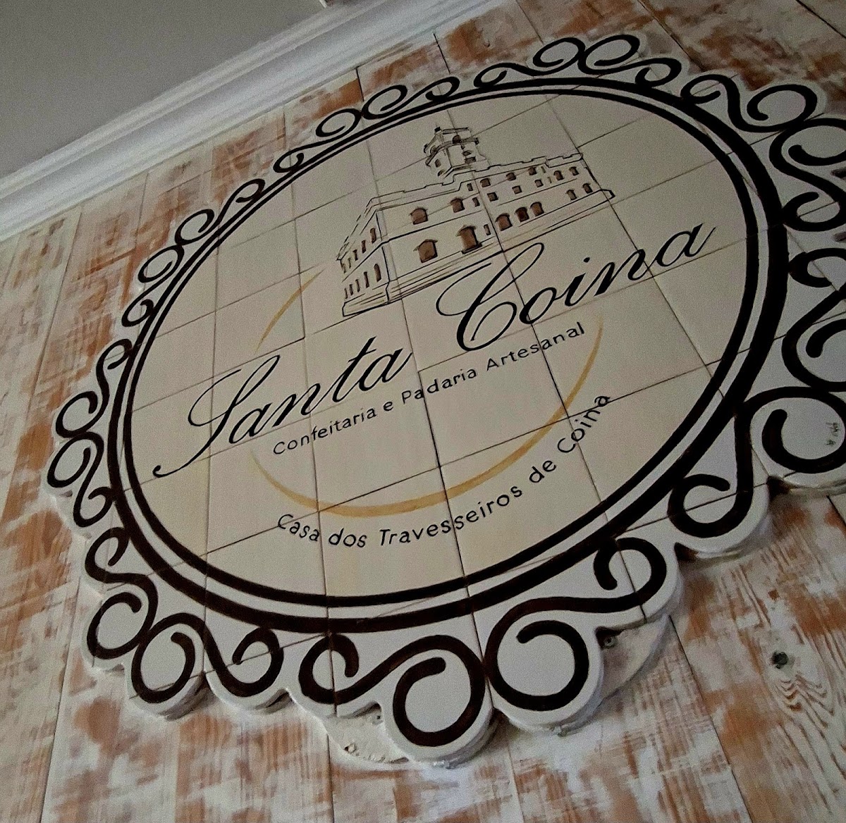 O emblema da Santa Coina pintado no chão da loja: o antigo edifício de Coina dentro de uma moldura de filigrana, sobre madeira branqueada.